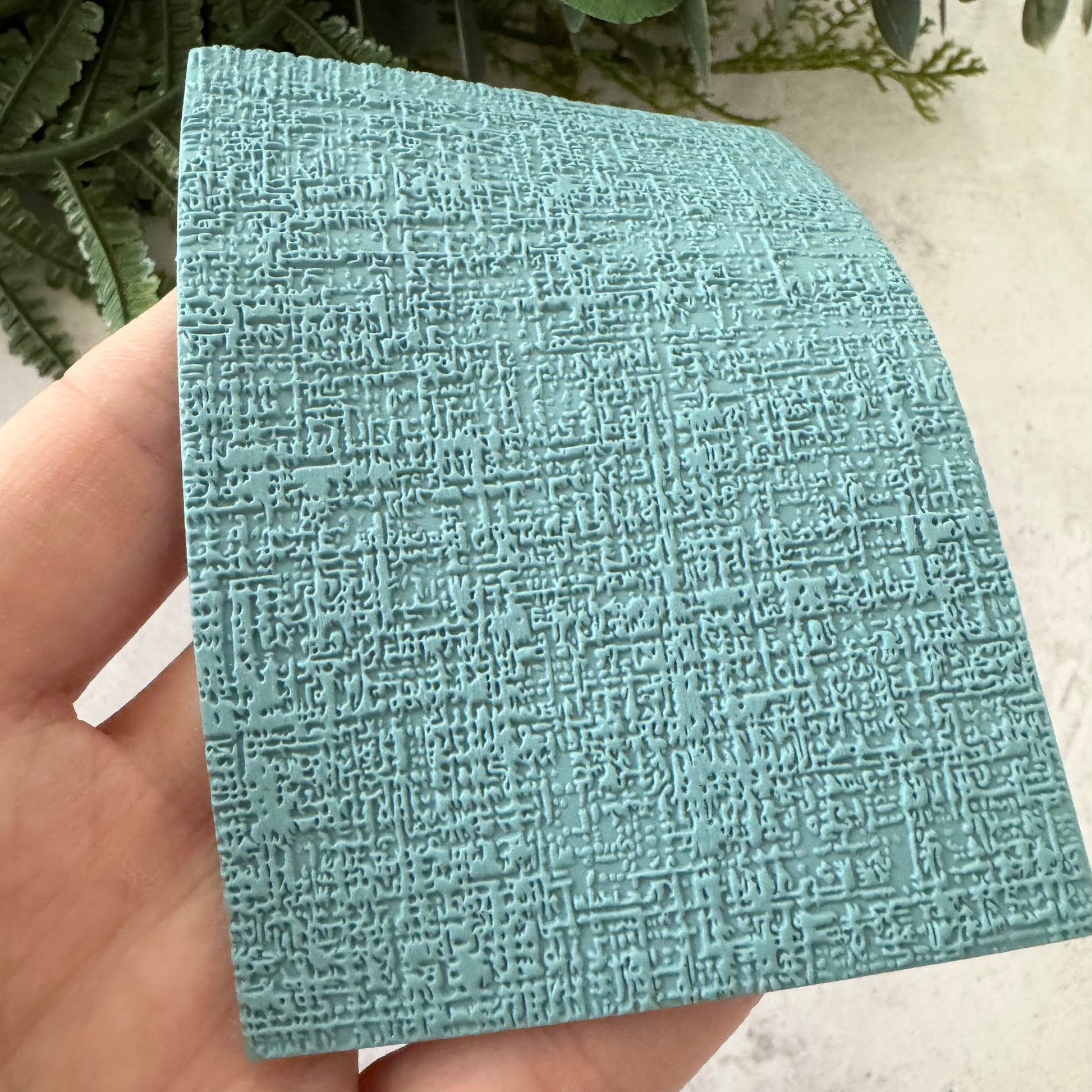 A hand holding a square sample of textured, embossed aqua blue material, showing its detailed pattern against a light background with green leaves.