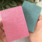 Two textured rectangular cards or small booklets held in a hand, one pink and one teal, with green leaves in the background.