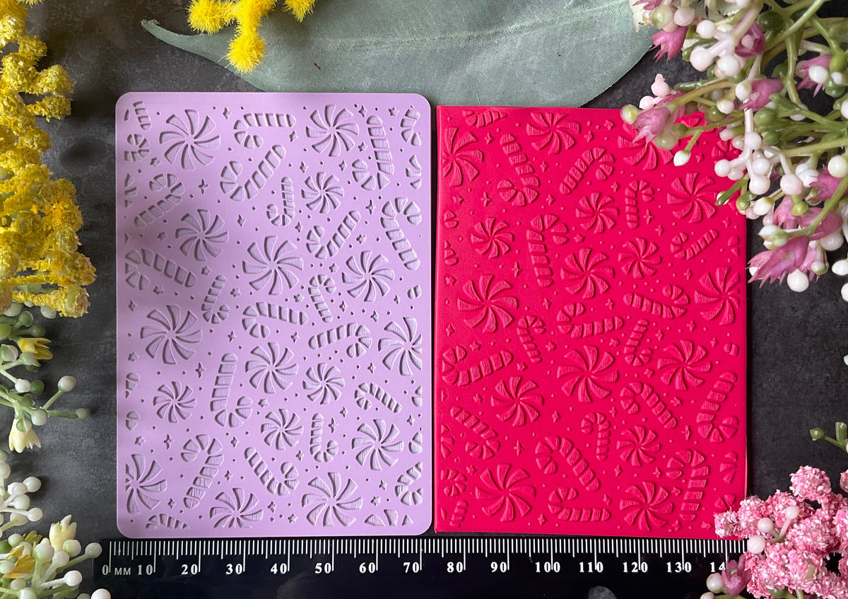 Two rectangular embossed stamp blocks, one light lavender and one bright pink, displayed side-by-side on a dark textured surface with peppermints and candy canes and stars pattern with a black ruler at the bottom and floral decorations around the edges.