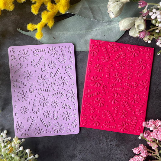 Two rectangular embossed stamping plates with a candy canes and peppermints and stars pattern, one light purple and one bright pink, displayed on a dark textured surface surrounded by yellow and pink flowers.