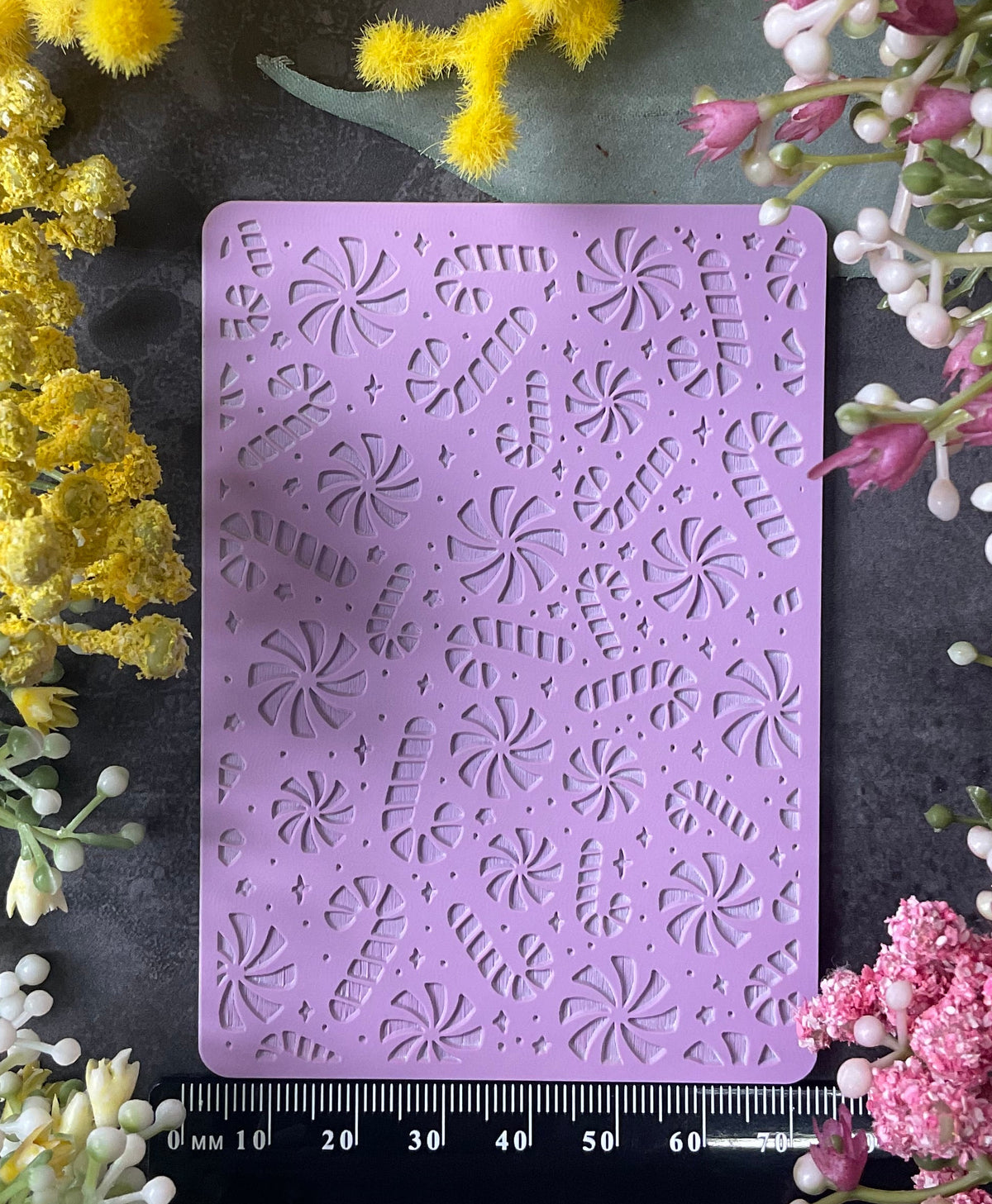 A lavender purple silicone stamping or embossing mat with a Christmas candy canes and peppermints and stars pattern, displayed on a dark gray surface and surrounded by colorful flowers (yellow, pink, white) with a black measuring ruler at the bottom.
