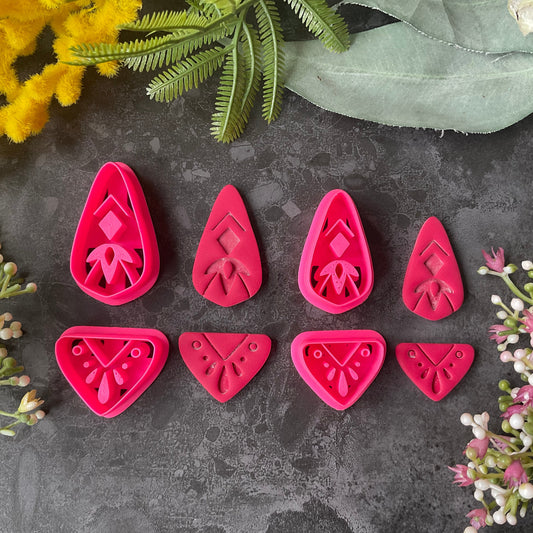 Bright pink 3d printed polymer clay cutters with floral and geometric designs are arranged on a dark textured surface with a black ruler at the bottom for scale, surrounded by decorative flowers and green leaves. Dune Drop Polymer Clay Cutter Set