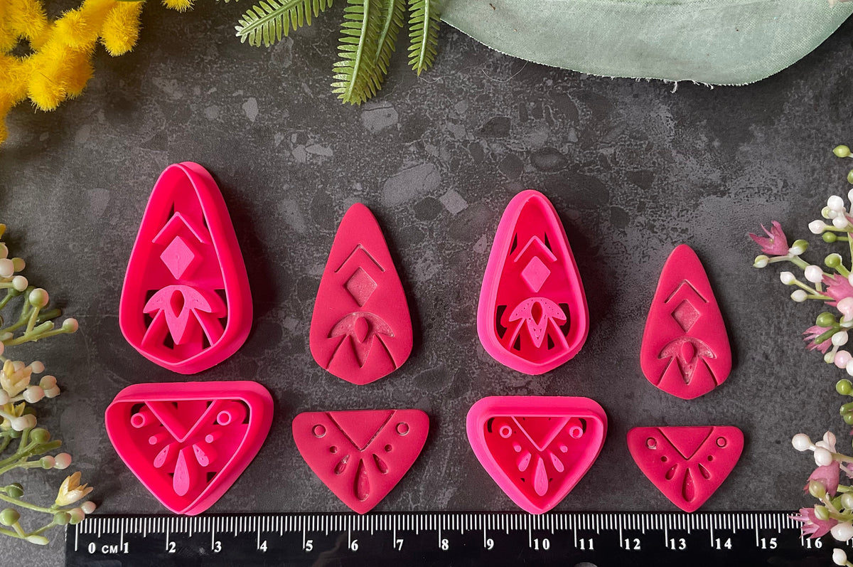 Bright pink 3d printed polymer clay cutters with floral and geometric designs are arranged on a dark textured surface with a black ruler at the bottom for scale, surrounded by decorative flowers and green leaves. Dune Drop Polymer Clay Cutter Set