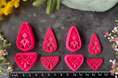 Bright pink 3d printed polymer clay cutters with floral and geometric designs are arranged on a dark textured surface with a black ruler at the bottom for scale, surrounded by decorative flowers and green leaves. Dune Drop Polymer Clay Cutter Set