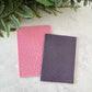 Two textured notebooks, one pink and one purple, on a light surface with green leaves in the background.