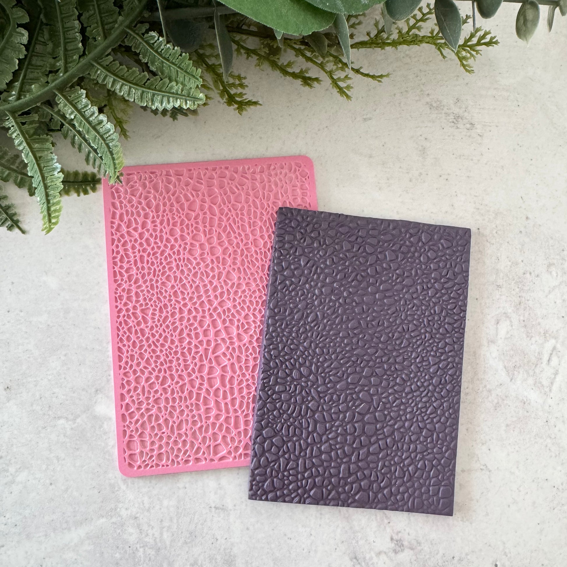 Two textured notebooks, one pink and one purple, on a light surface with green leaves in the background.