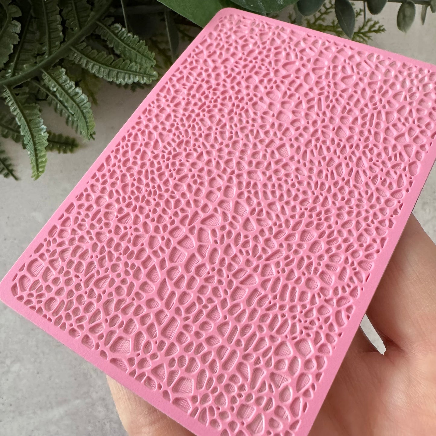 A pink rectangular silicone mold or stamp with a textured, raised organic pattern, held in a hand against a background of green plants and a light gray surface.