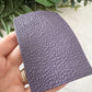 A hand holding a textured, pebbled faux leather or vinyl material sample in a muted purple color, with green leaves in the background.