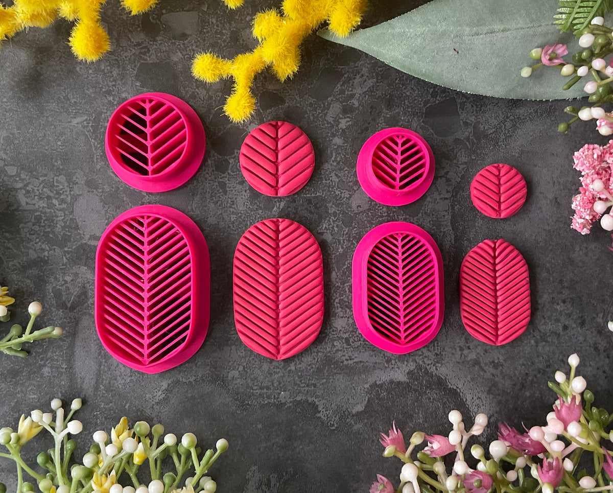 A set of bright pink 3d printed leaf-shaped polymer clay cutters arranged on a dark gray textured surface, surrounded by yellow and pink flowers. Etched Forms Polymer Clay Cutter Set