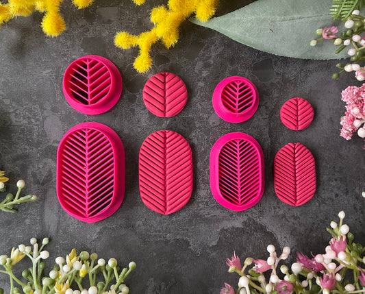 A set of bright pink 3d printed leaf-shaped polymer clay cutters arranged on a dark gray textured surface, surrounded by yellow and pink flowers. Etched Forms Polymer Clay Cutter Set
