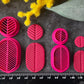 A set of bright pink 3d printed leaf-shaped polymer clay cutters arranged on a dark gray textured surface, surrounded by yellow and pink flowers. Etched Forms Polymer Clay Cutter Set