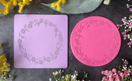 A flat-lay product photo showing a lavender rectangular mold with an embossed Eucalyptus flowers and gumnuts floral wreath design next to a pink circular mold with a matching floral border, styled with yellow flowers and green leaves on a dark gray background.
