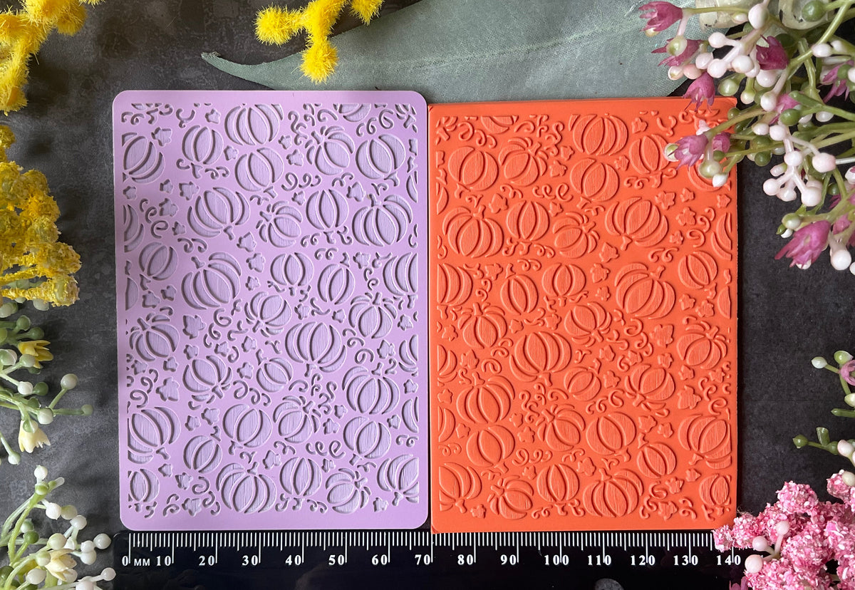 Two rectangular embossed craft mats or cards in lavender and coral orange with fall Halloween pumpkin and leaves texture, laid side by side on a dark surface with a black ruler underneath and yellow and pink flowers around the edges.
