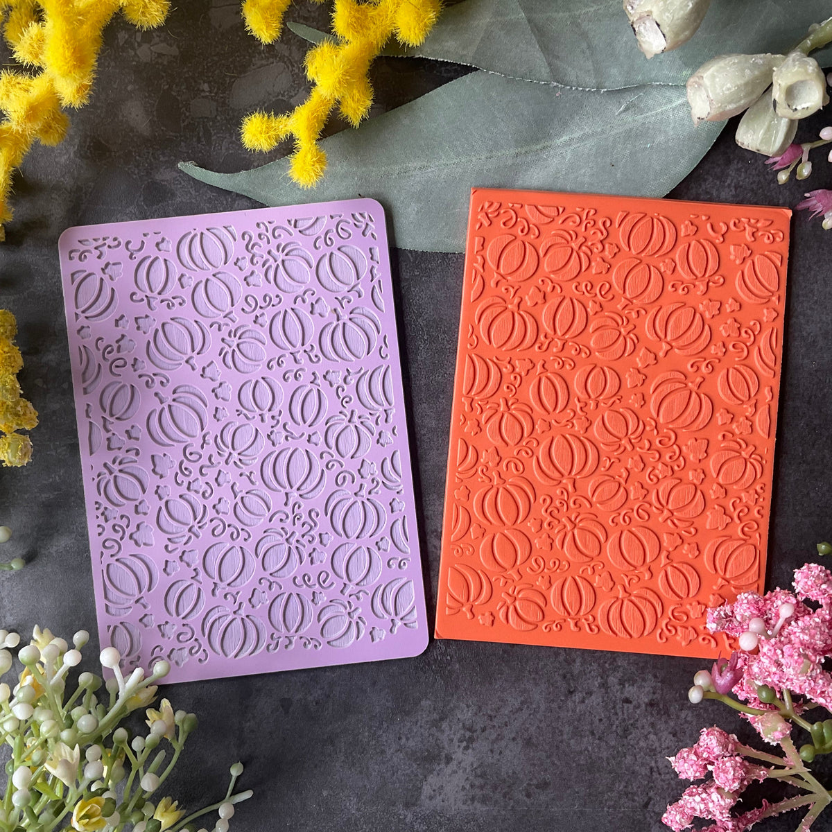 Two rectangular embossed stamp blocks or cards in lavender purple and bright orange with fall Halloween pumpkins and leaves texture, arranged on a dark textured surface with yellow and pink flowers around them.
