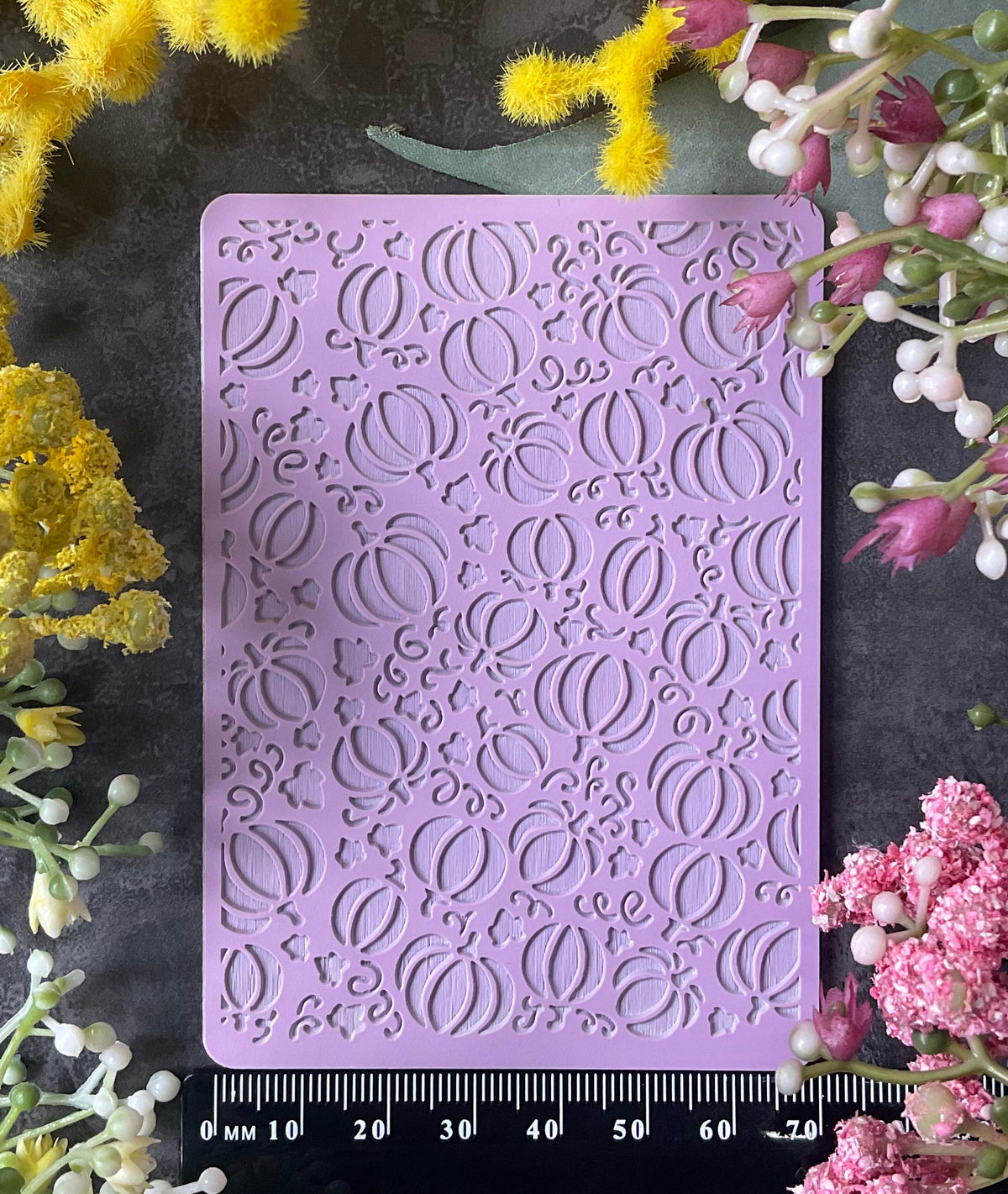 A lavender purple rectangular embossed stamp or mold with a repeating fall Halloween pumpkin and leaves pattern, displayed on a dark textured surface and surrounded by yellow and pink flowers. A black ruler is placed underneath the item for scale.