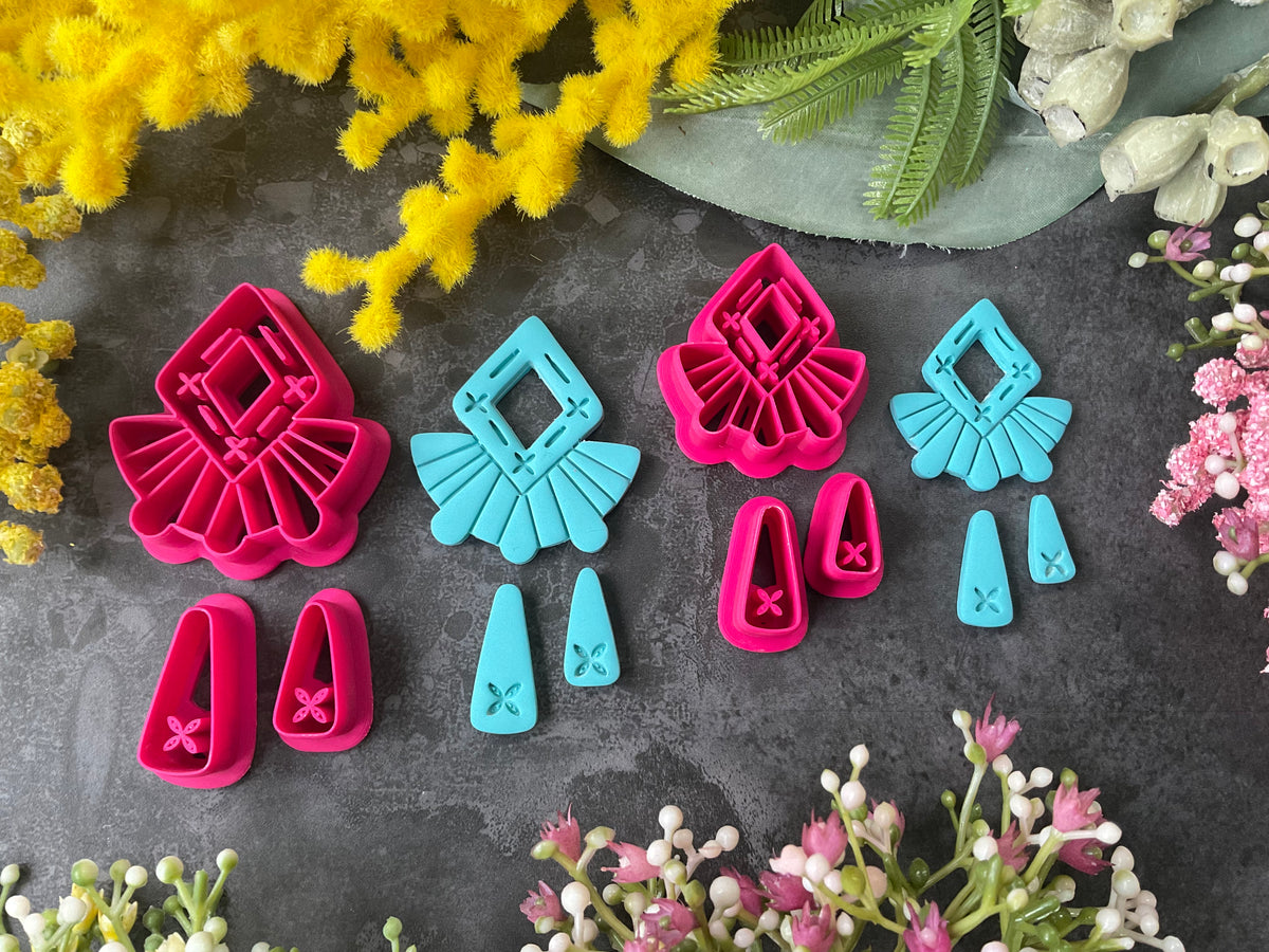 A set of bright pink and turquoise 3d printed polymer clay cutters arranged on a dark textured surface with decorative flowers around the edges. The set includes large fan-shaped stamping cutters, smaller matching cutters, and pointed cone-shaped cutters. First Light Polymer Clay Cutter Set