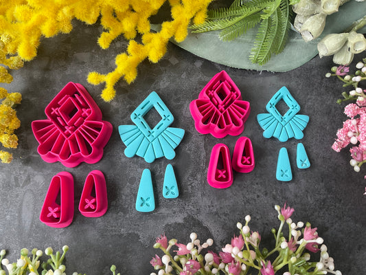 A set of bright pink and turquoise 3d printed polymer clay cutters arranged on a dark textured surface with decorative flowers around the edges. The set includes large fan-shaped stamping cutters, smaller matching cutters, and pointed cone-shaped cutters. First Light Polymer Clay Cutter Set