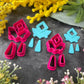 A set of bright pink and turquoise 3d printed polymer clay cutters arranged on a dark textured surface with decorative flowers around the edges. The set includes large fan-shaped stamping cutters, smaller matching cutters, and pointed cone-shaped cutters. First Light Polymer Clay Cutter Set