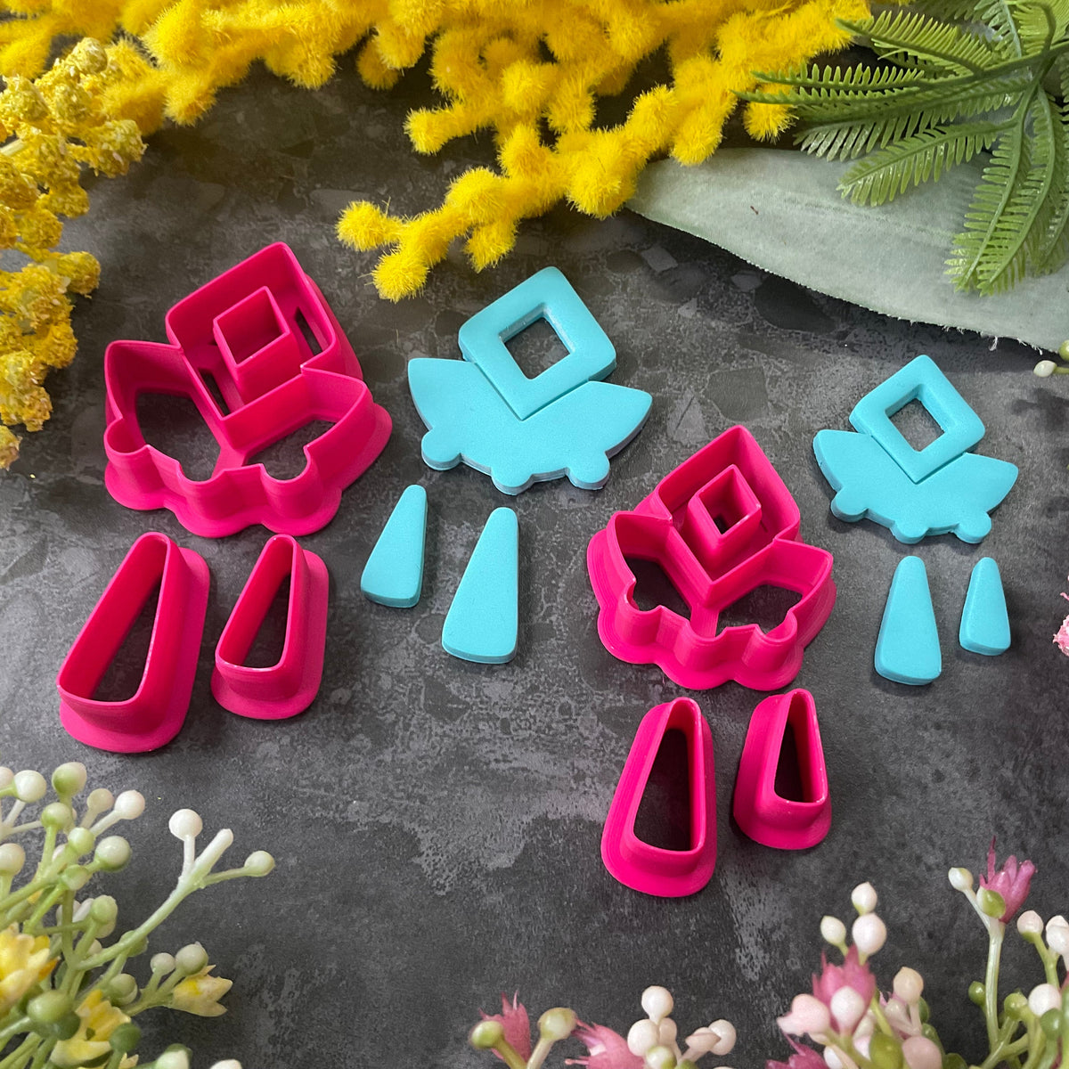 A set of bright pink and turquoise 3d printed polymer clay cutters arranged on a dark textured surface with decorative flowers around the edges. The set includes large fan-shaped stamping cutters, smaller matching cutters, and pointed cone-shaped cutters. First Light Polymer Clay Cutter Set