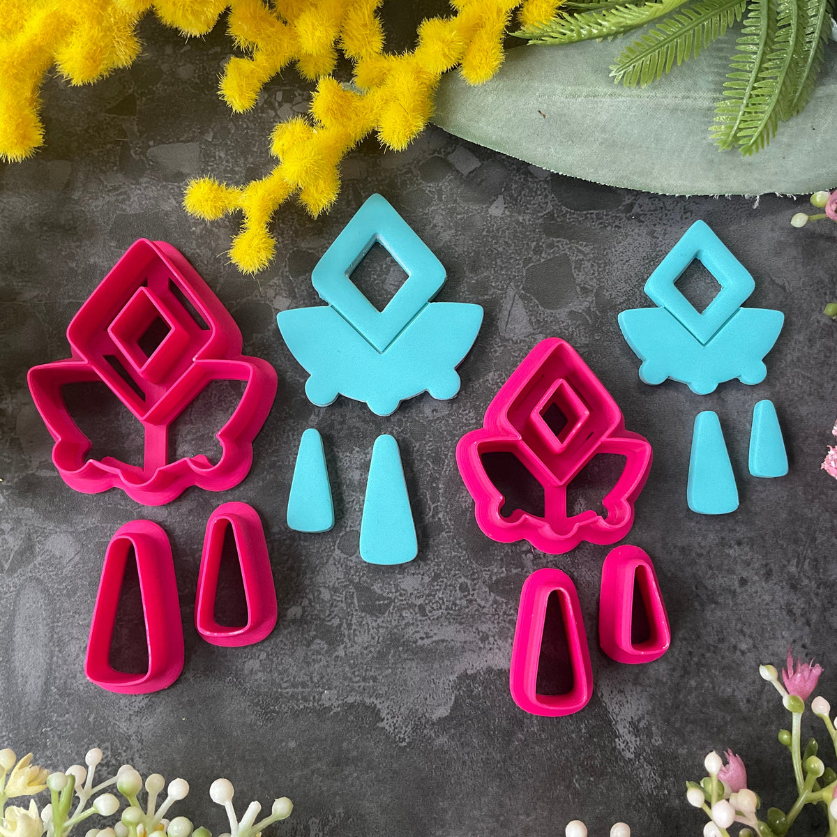 A set of bright pink and turquoise 3d printed polymer clay cutters arranged on a dark textured surface with decorative flowers around the edges. The set includes large fan-shaped stamping cutters, smaller matching cutters, and pointed cone-shaped cutters. First Light Polymer Clay Cutter Set