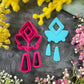A set of bright pink and turquoise 3d printed polymer clay cutters arranged on a dark textured surface with decorative flowers around the edges. The set includes large fan-shaped stamping cutters, smaller matching cutters, and pointed cone-shaped cutters. First Light Polymer Clay Cutter Set