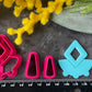 A set of bright pink and turquoise 3d printed polymer clay cutters arranged on a dark textured surface with decorative flowers around the edges. The set includes large fan-shaped stamping cutters, smaller matching cutters, and pointed cone-shaped cutters. First Light Polymer Clay Cutter Set
