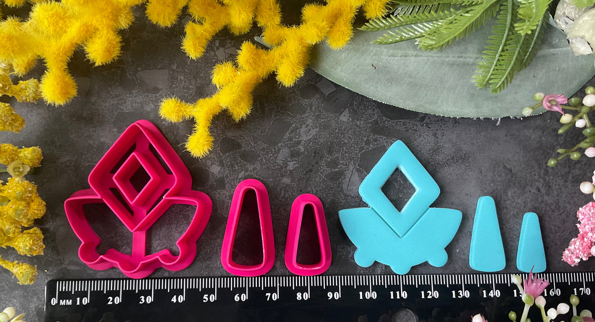 A set of bright pink and turquoise 3d printed polymer clay cutters arranged on a dark textured surface with decorative flowers around the edges. The set includes large fan-shaped stamping cutters, smaller matching cutters, and pointed cone-shaped cutters. First Light Polymer Clay Cutter Set