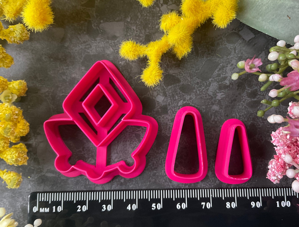 A set of bright pink and turquoise 3d printed polymer clay cutters arranged on a dark textured surface with decorative flowers around the edges. The set includes large fan-shaped stamping cutters, smaller matching cutters, and pointed cone-shaped cutters. First Light Polymer Clay Cutter Set
