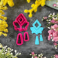 A set of bright pink and turquoise 3d printed polymer clay cutters arranged on a dark textured surface with decorative flowers around the edges. The set includes large fan-shaped stamping cutters, smaller matching cutters, and pointed cone-shaped cutters. First Light Polymer Clay Cutter Set