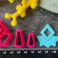 A set of bright pink and turquoise 3d printed polymer clay cutters arranged on a dark textured surface with decorative flowers around the edges. The set includes large fan-shaped stamping cutters, smaller matching cutters, and pointed cone-shaped cutters. First Light Polymer Clay Cutter Set