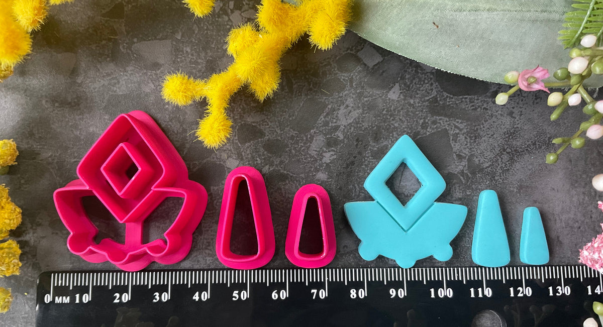 A set of bright pink and turquoise 3d printed polymer clay cutters arranged on a dark textured surface with decorative flowers around the edges. The set includes large fan-shaped stamping cutters, smaller matching cutters, and pointed cone-shaped cutters. First Light Polymer Clay Cutter Set