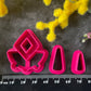A set of bright pink and turquoise 3d printed polymer clay cutters arranged on a dark textured surface with decorative flowers around the edges. The set includes large fan-shaped stamping cutters, smaller matching cutters, and pointed cone-shaped cutters. First Light Polymer Clay Cutter Set