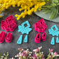 A set of bright pink and turquoise 3d printed polymer clay cutters arranged on a dark textured surface with decorative flowers around the edges. The set includes large fan-shaped stamping cutters, smaller matching cutters, and pointed cone-shaped cutters. First Light Polymer Clay Cutter Set