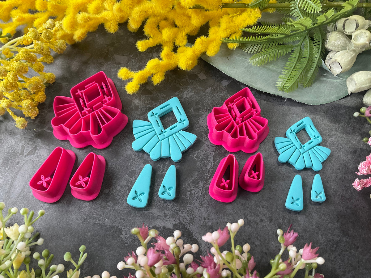 A set of bright pink and turquoise 3d printed polymer clay cutters arranged on a dark textured surface with decorative flowers around the edges. The set includes large fan-shaped stamping cutters, smaller matching cutters, and pointed cone-shaped cutters. First Light Polymer Clay Cutter Set