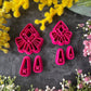 A set of bright pink and turquoise 3d printed polymer clay cutters arranged on a dark textured surface with decorative flowers around the edges. The set includes large fan-shaped stamping cutters, smaller matching cutters, and pointed cone-shaped cutters. First Light Polymer Clay Cutter Set