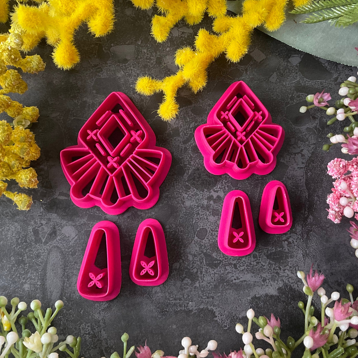 A set of bright pink and turquoise 3d printed polymer clay cutters arranged on a dark textured surface with decorative flowers around the edges. The set includes large fan-shaped stamping cutters, smaller matching cutters, and pointed cone-shaped cutters. First Light Polymer Clay Cutter Set