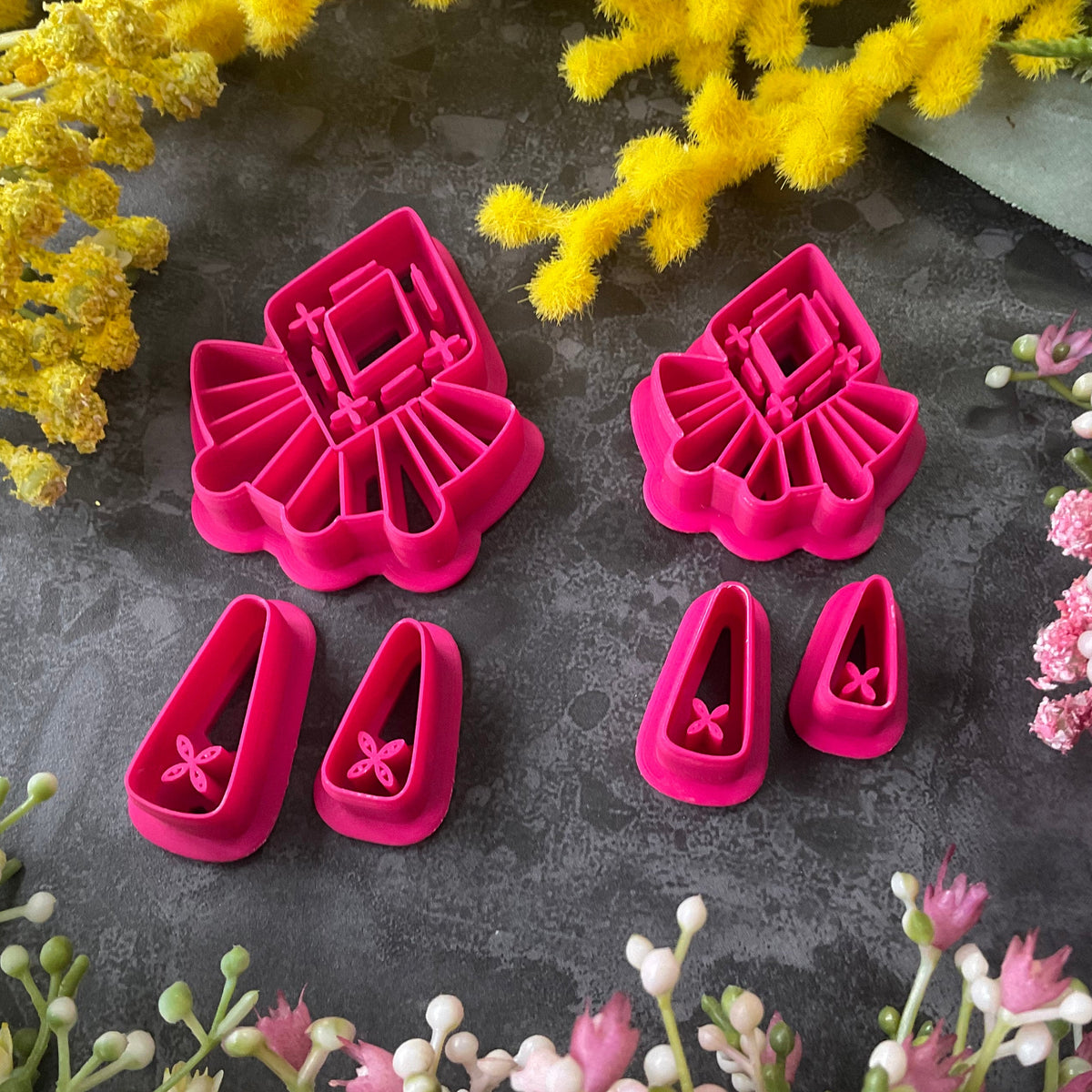 A set of bright pink and turquoise 3d printed polymer clay cutters arranged on a dark textured surface with decorative flowers around the edges. The set includes large fan-shaped stamping cutters, smaller matching cutters, and pointed cone-shaped cutters. First Light Polymer Clay Cutter Set