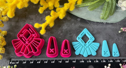 A set of bright pink and turquoise 3d printed polymer clay cutters arranged on a dark textured surface with decorative flowers around the edges. The set includes large fan-shaped stamping cutters, smaller matching cutters, and pointed cone-shaped cutters. First Light Polymer Clay Cutter Set
