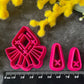 A set of bright pink and turquoise 3d printed polymer clay cutters arranged on a dark textured surface with decorative flowers around the edges. The set includes large fan-shaped stamping cutters, smaller matching cutters, and pointed cone-shaped cutters. First Light Polymer Clay Cutter Set