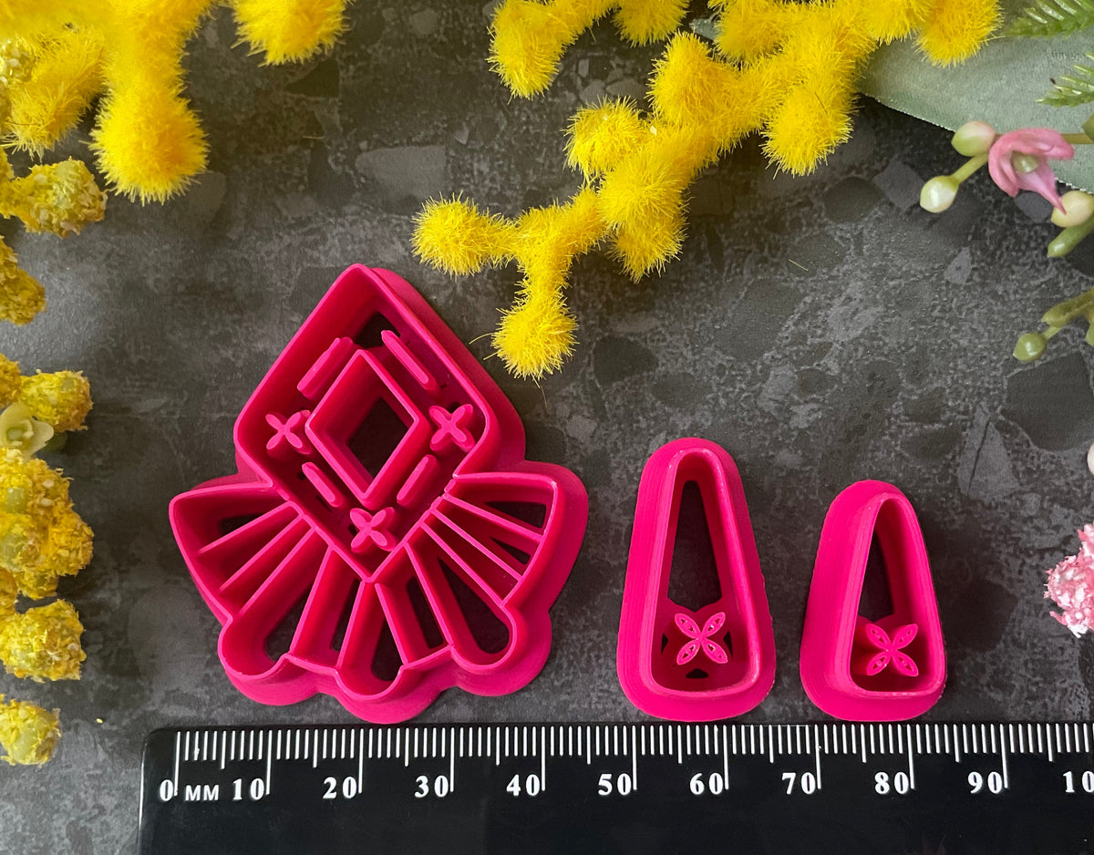 A set of bright pink and turquoise 3d printed polymer clay cutters arranged on a dark textured surface with decorative flowers around the edges. The set includes large fan-shaped stamping cutters, smaller matching cutters, and pointed cone-shaped cutters. First Light Polymer Clay Cutter Set