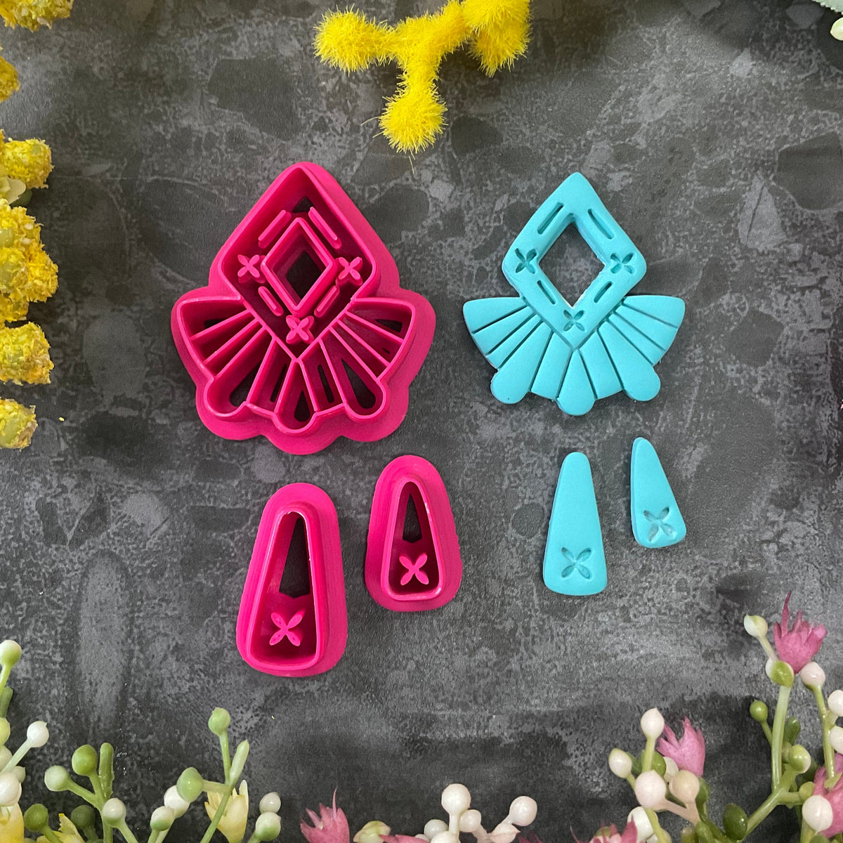A set of bright pink and turquoise 3d printed polymer clay cutters arranged on a dark textured surface with decorative flowers around the edges. The set includes large fan-shaped stamping cutters, smaller matching cutters, and pointed cone-shaped cutters. First Light Polymer Clay Cutter Set