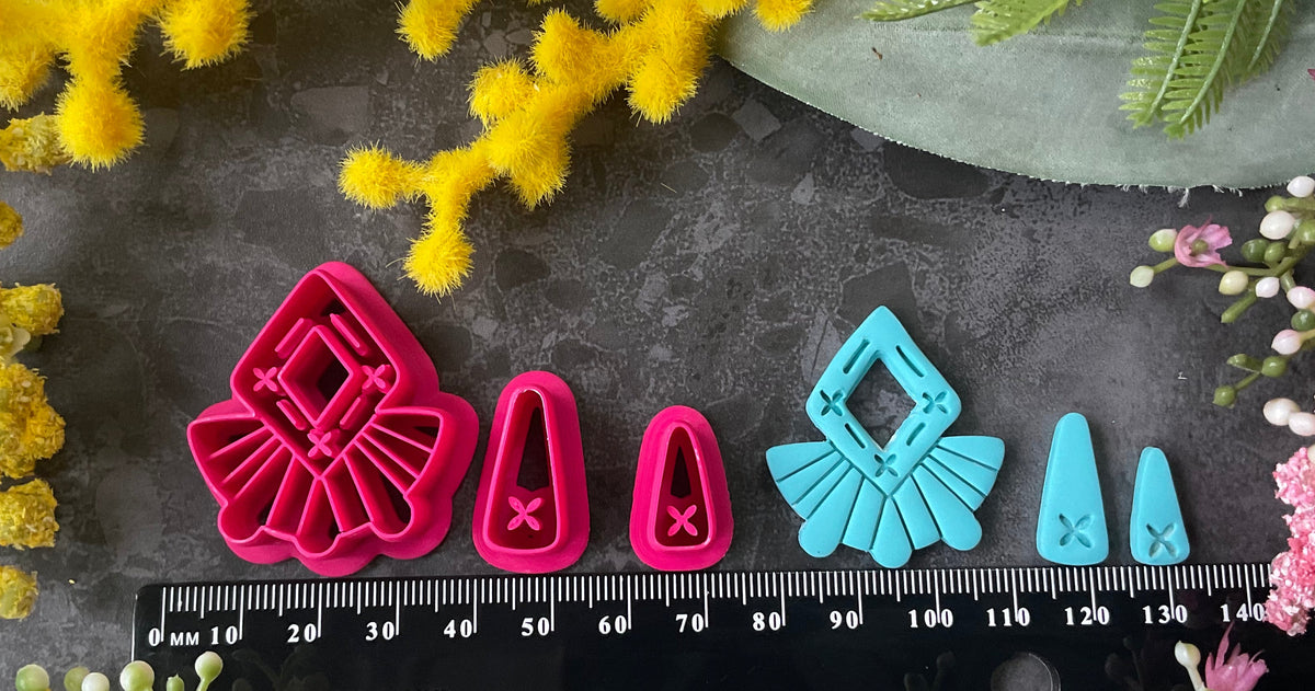 A set of bright pink and turquoise 3d printed polymer clay cutters arranged on a dark textured surface with decorative flowers around the edges. The set includes large fan-shaped stamping cutters, smaller matching cutters, and pointed cone-shaped cutters. First Light Polymer Clay Cutter Set
