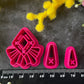 A set of bright pink and turquoise 3d printed polymer clay cutters arranged on a dark textured surface with decorative flowers around the edges. The set includes large fan-shaped stamping cutters, smaller matching cutters, and pointed cone-shaped cutters. First Light Polymer Clay Cutter Set