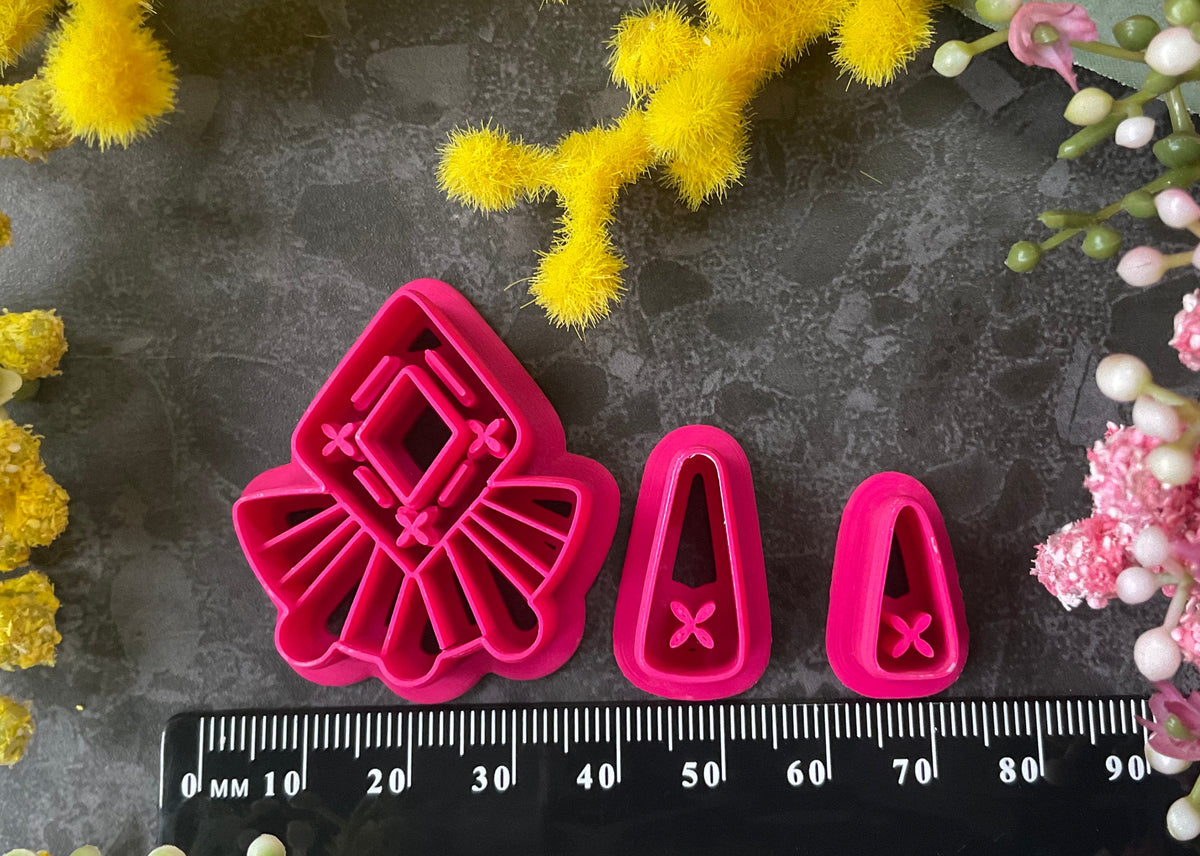 A set of bright pink and turquoise 3d printed polymer clay cutters arranged on a dark textured surface with decorative flowers around the edges. The set includes large fan-shaped stamping cutters, smaller matching cutters, and pointed cone-shaped cutters. First Light Polymer Clay Cutter Set