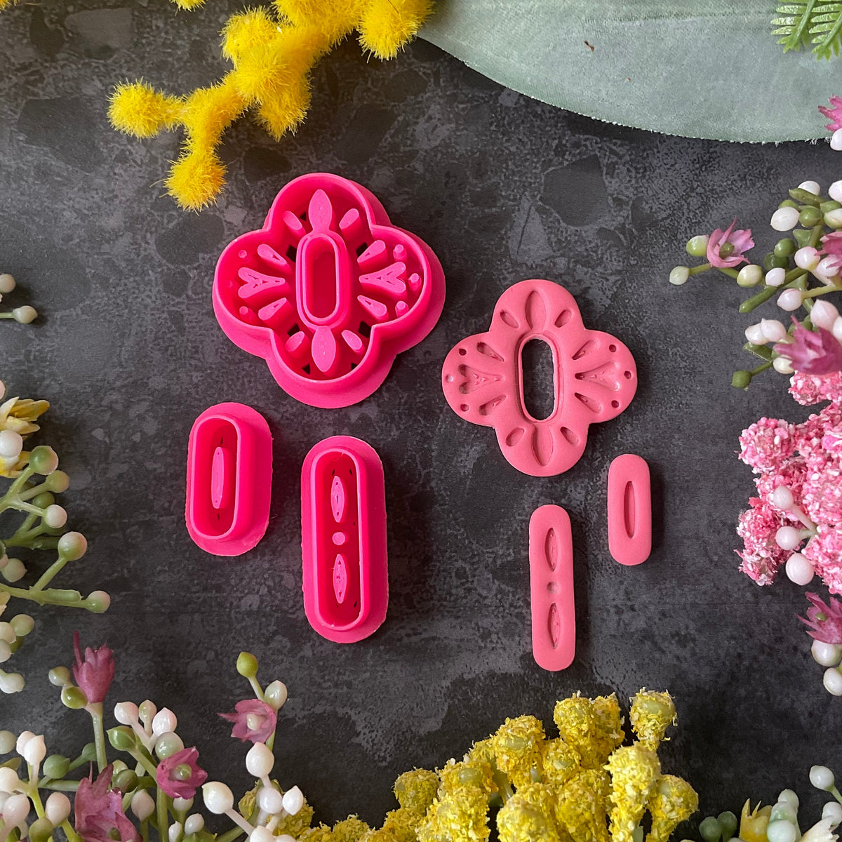 A set of bright pink 3d printed polymer clay cutters arranged on a dark textured surface, surrounded by decorative flowers in yellow, pink, and white. The cutters are a Mediterranean tile style design, Folk Bloom Polymer Clay Cutter Set.