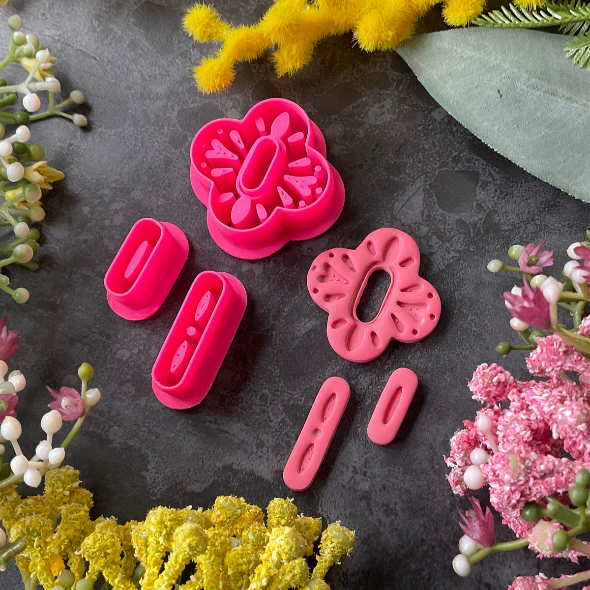 A set of bright pink 3d printed polymer clay cutters arranged on a dark textured surface, surrounded by decorative flowers in yellow, pink, and white. The cutters are a Mediterranean tile style design, Folk Bloom Polymer Clay Cutter Set.