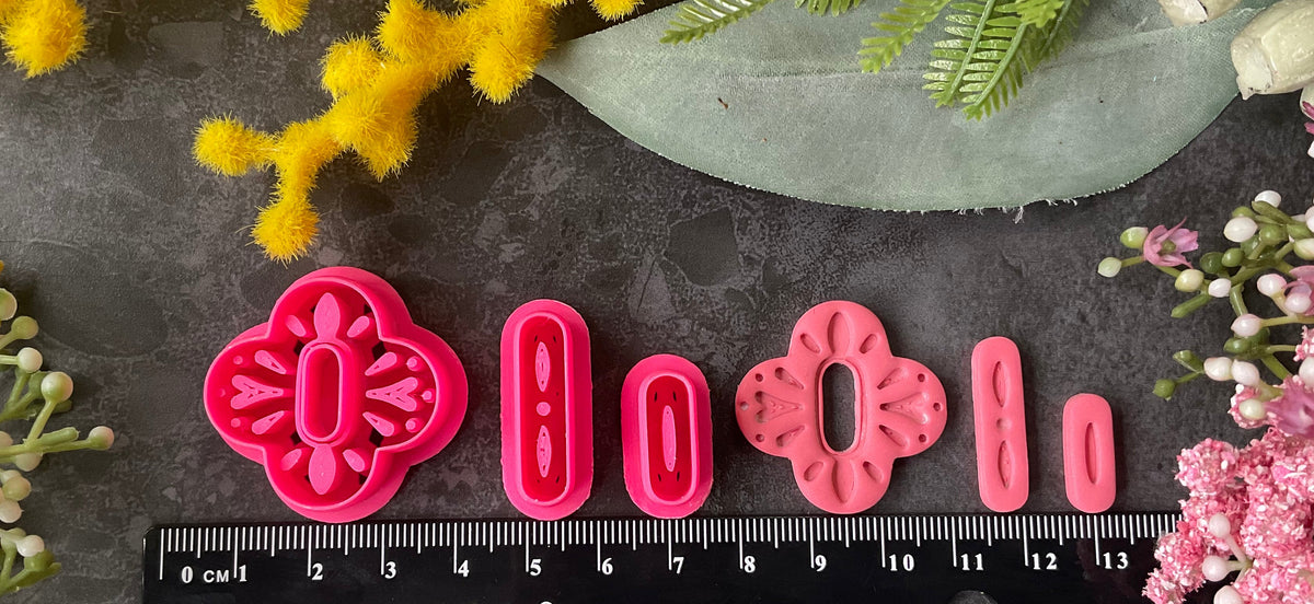 A set of bright pink 3d printed polymer clay cutters arranged on a dark textured surface, surrounded by decorative flowers in yellow, pink, and white. The cutters are a Mediterranean tile style design, Folk Bloom Polymer Clay Cutter Set.