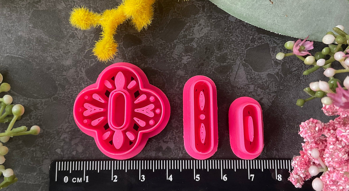 A set of bright pink 3d printed polymer clay cutters arranged on a dark textured surface, surrounded by decorative flowers in yellow, pink, and white. The cutters are a Mediterranean tile style design, Folk Bloom Polymer Clay Cutter Set.