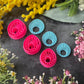 Three 3d printed polymer clay cutters in bright pink, arranged on a dark textured surface surrounded by colorful flowers. Halo Beam Polymer Clay Cutter