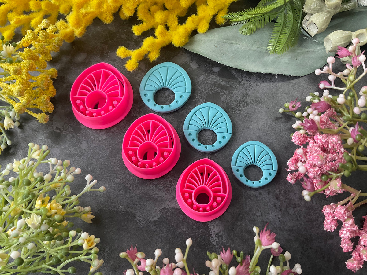 Three 3d printed polymer clay cutters in bright pink, arranged on a dark textured surface surrounded by colorful flowers. Halo Beam Polymer Clay Cutter